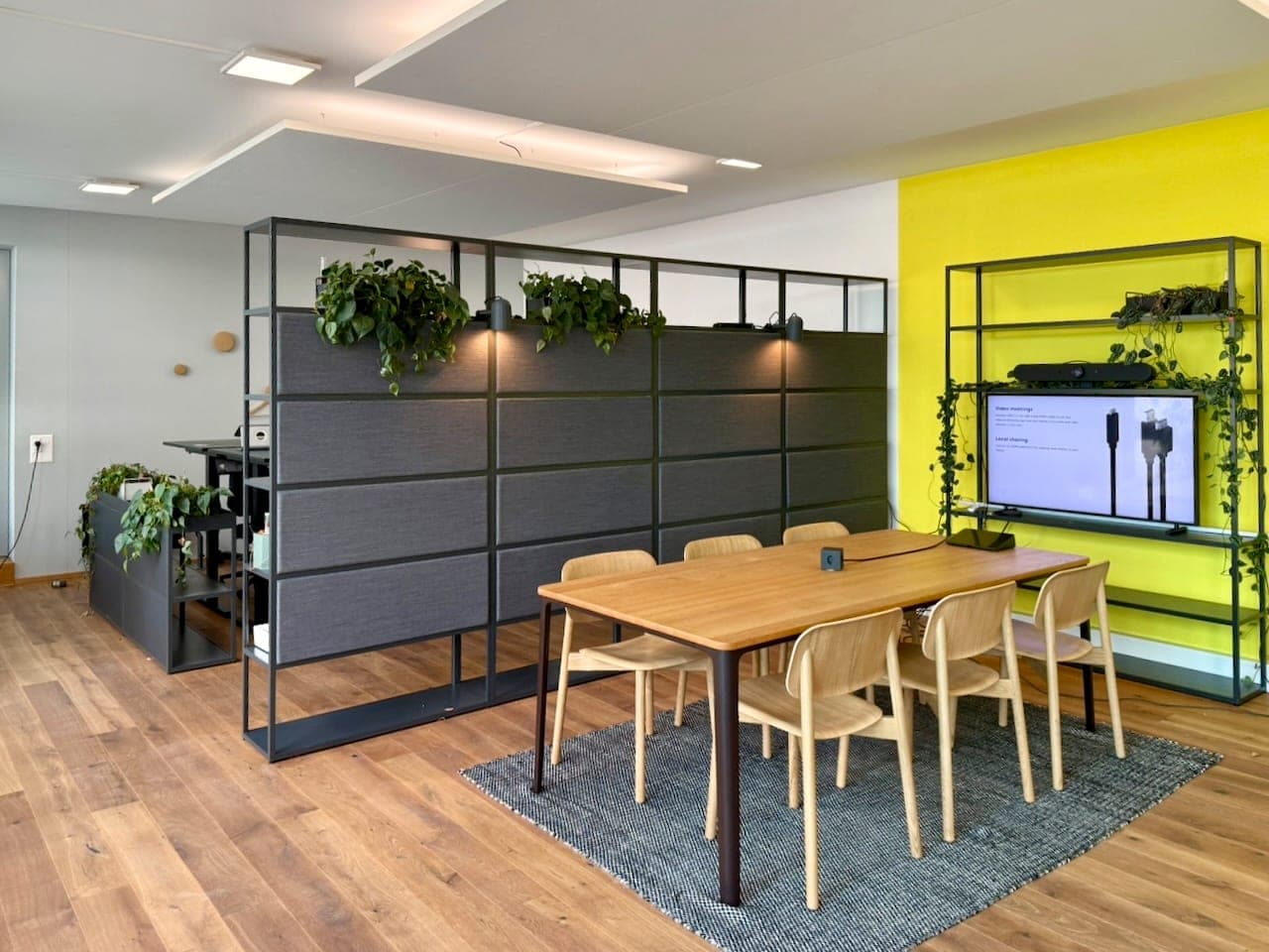 Komeo Corner in Luzern: Holztisch mit sechs Stühlen vor gelber Wand mit Screen, dahinter Trennregal