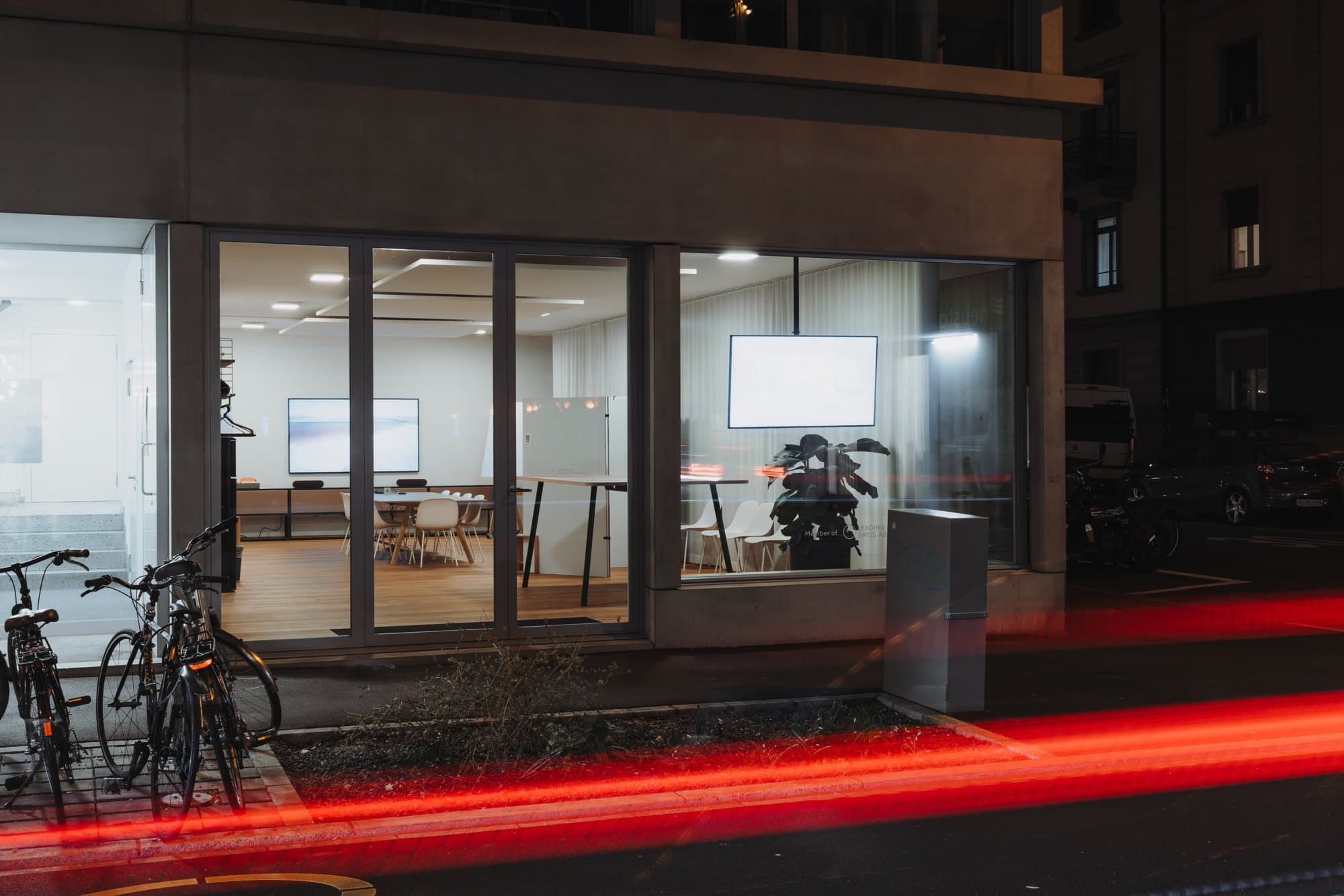 Das Erdgeschoss an der Neustadtstrasse am Abend: hell erleuchtete Räume hinter der Glasfront, davor Velos und die Lichtspur eines Autos
