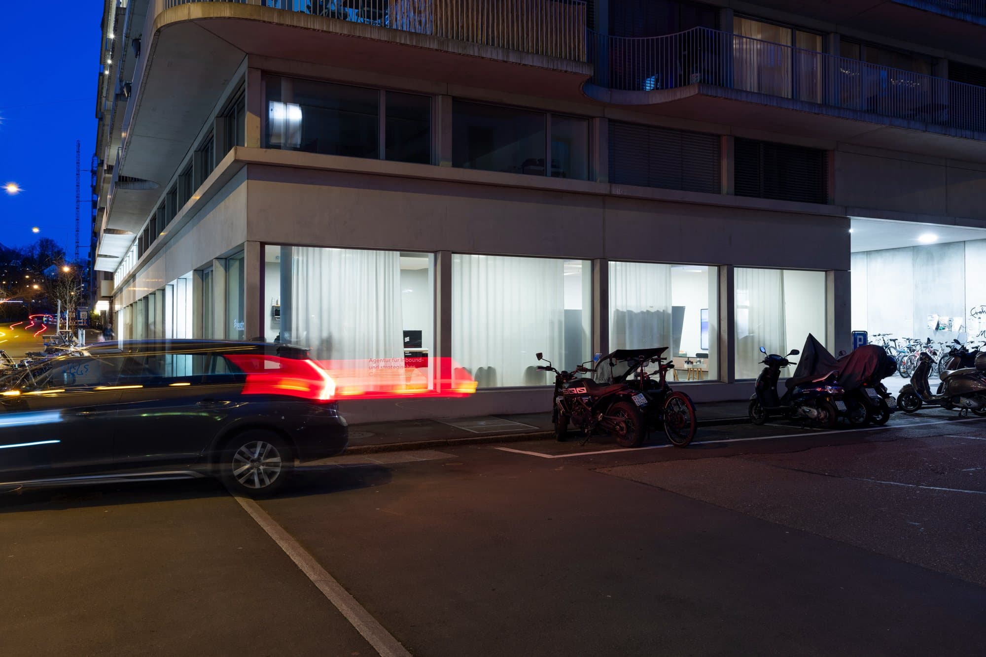 Die beleuchtete Fensterfront im Erdgeschoss an der Strassenecke, am Abend